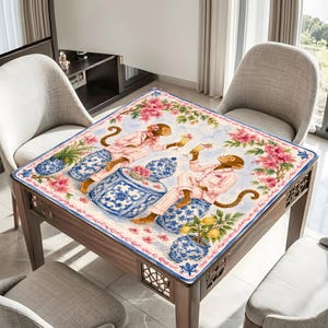 May include: Square wooden table with a decorative cloth featuring a whimsical design. The cloth displays monkeys in pink pajamas, blue and white vases, and pink floral accents. The table is surrounded by chairs.