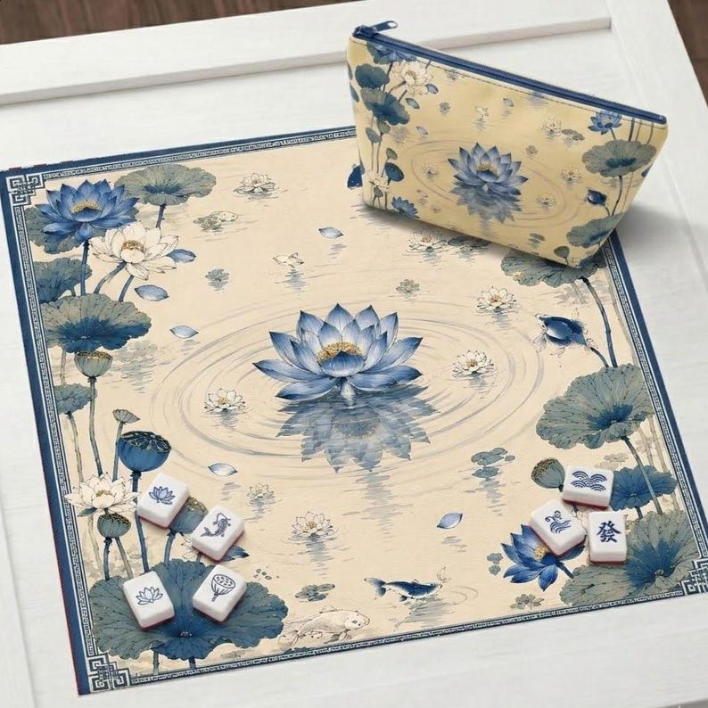 May include: A square mahjong mat and a matching zippered pouch, both featuring a cream background with blue lotus flowers, lily pads, and ripples. Several white mahjong tiles with blue ink designs are scattered across the mat surface.