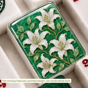 Pu&ograve; includere: Una piastrella rettangolare bianca con un disegno stampato di quattro fiori di giglio bianchi con stami gialli e foglie verdi su uno sfondo verde smeraldo testurizzato. La piastrella ha una finitura lucida e bordi arrotondati.