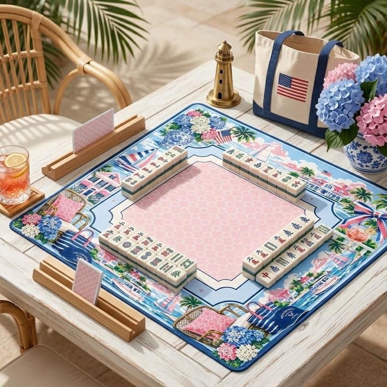 May include: A mahjong set arranged on a table, featuring a light blue and pink game mat with nautical and floral designs. The set includes tiles, racks, and a tote bag with an American flag. A gold lighthouse figurine and a vase of hydrangeas are also present.