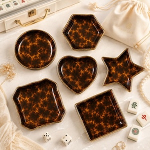 Puede incluir: Un conjunto de seis platos pequeños de diversas formas, incluyendo un círculo, un hexágono, un corazón, una estrella y dos cuadrados. Cada plato presenta un patrón de carey en tonos marrón oscuro y naranja con acabado brillante y bordes dorados.