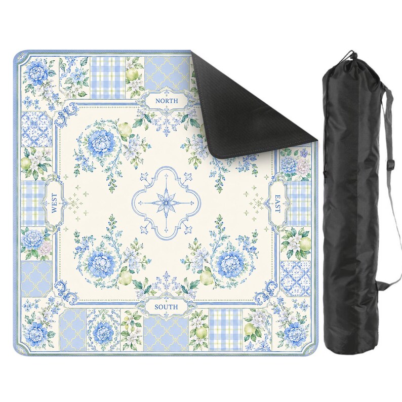 May include: A square picnic blanket with a floral and plaid pattern in shades of blue, white, and green. The blanket has a compass rose design in the center and is labeled with the cardinal directions. A black carrying bag is included.