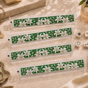 Pu&ograve; includere: Quattro supporti in acrilico trasparente per tessere del mahjong con un inserto decorativo caratterizzato da un motivo ripetuto di fiori di giglio bianchi e foglie verdi su sfondo verde scuro. Ogni supporto ha una piccola vite nera a sinistra.