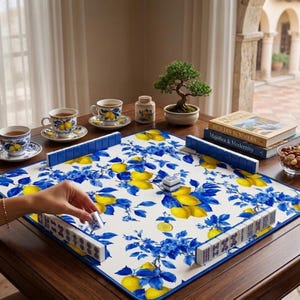 May include: A mahjong set with a white, blue, and yellow floral design, featuring lemons and blue leaves. The set is arranged on a dark wooden table, with teacups, saucers, and a bonsai tree in the background. A hand is placing a tile.