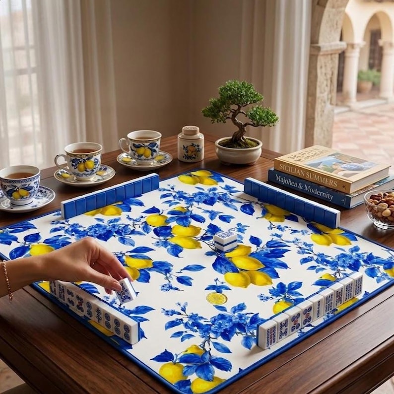 May include: A mahjong set with a white, blue, and yellow floral design, featuring lemons and blue leaves. The set is arranged on a dark wooden table, with teacups, saucers, and a bonsai tree in the background. A hand is placing a tile.