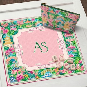 Op de afbeelding: Een roze en groene Mahjong-spelset met een bijpassend etui met ritssluiting. Het spelbord heeft een roze midden met de initialen "AS" en is omgeven door een kleurrijk bloemen- en pagodedesign. Er zijn verschillende Mahjong-tegels zichtbaar.