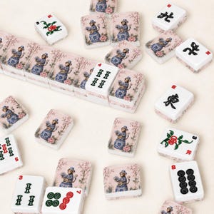 Op de afbeelding: Een verzameling witte Mahjong-tegels met roze bloemen- en apenontwerpen. Sommige tegels tonen Chinese karakters en stippen in zwart en rood. De tegels zijn verspreid over een lichtgekleurd oppervlak.
