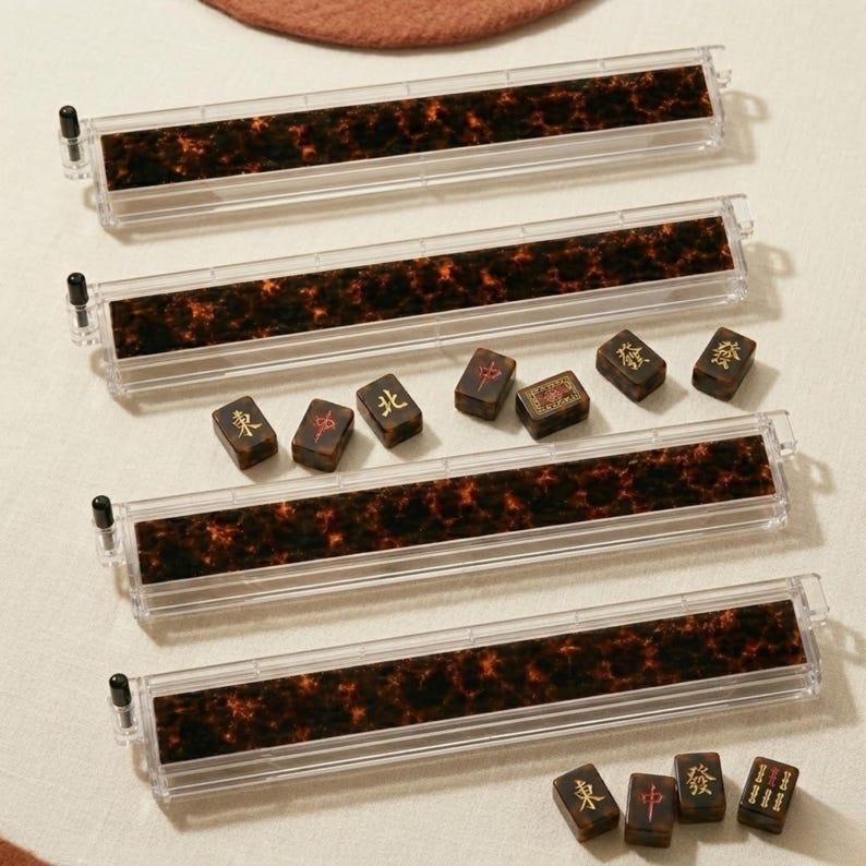 Puede incluir: Cuatro soportes rectangulares para fichas de mahjong hechos de acr&iacute;lico transparente con un inserto de patr&oacute;n de carey en marr&oacute;n oscuro y negro. Cada soporte cuenta con una peque&ntilde;a tapa negra en el extremo. Varias fichas de mahjong con caracteres dorados y rojos est&aacute;n esparcidas.