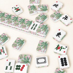 May include: A collection of white Mahjong tiles with various symbols and palm tree designs. Some tiles feature red roses. The tiles are scattered on a light beige surface, creating a visually appealing arrangement.