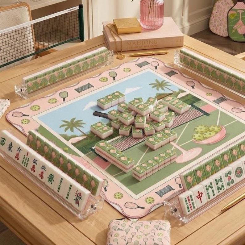 May include: A complete Mahjong set with green and white tiles, a pink and green playing mat with a tennis court design, and clear tile racks. The set is arranged on a light wood table, with a net and decorative items in the background.