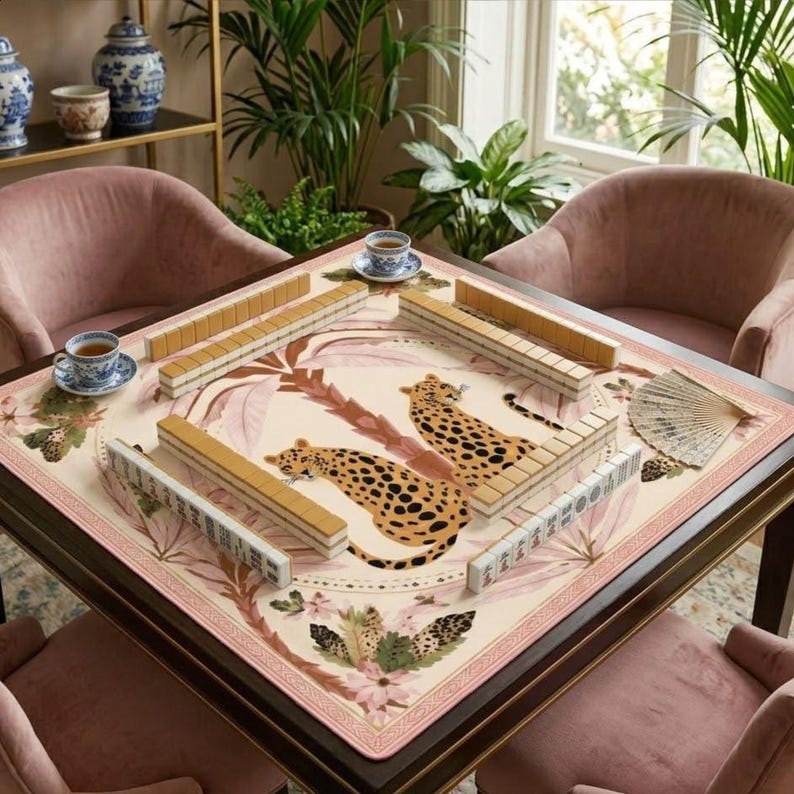 May include: A square mahjong table with a cream-colored playing surface featuring a leopard design and floral accents. The table is surrounded by pink armchairs. Tea cups and a fan are also visible.