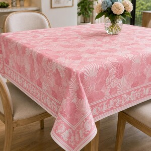 May include: A square tablecloth featuring a repeating pattern of white seashells and coral motifs on a soft pink background. The fabric has a distinct border design with intricate shell illustrations and a clean, straight-edged finish.