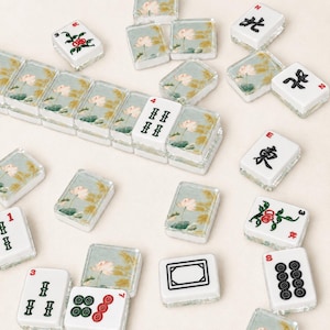 May include: A selection of white and clear Mahjong tiles, each with unique symbols and illustrations. Some tiles showcase floral patterns, while others display numbers and Chinese characters. The tiles are arranged on a light-coloured surface.