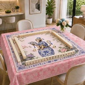 May include: A square mahjong mat featuring a light pink background with a central illustration of a monkey wearing a blue patterned outfit and holding a fan, seated on a blue and white porcelain jar, surrounded by mahjong tiles and floral motifs.