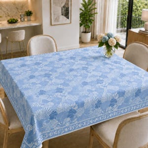 May include: A rectangular tablecloth featuring a repeating pattern of white seashells on a light blue background. The fabric has a distinct border design with a continuous seashell motif, covering a dining table surrounded by four upholstered chairs.