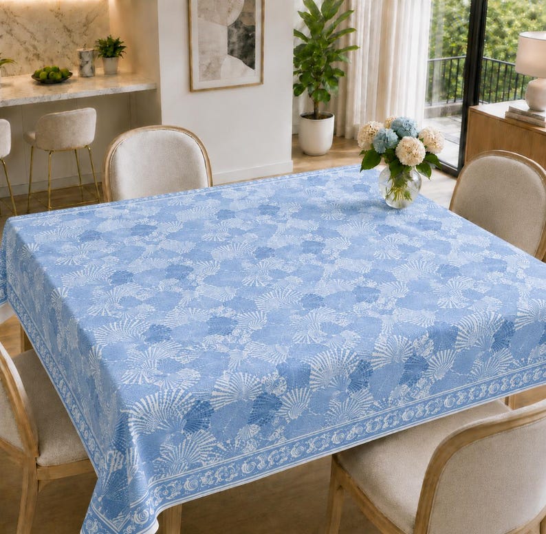 May include: A rectangular tablecloth featuring a repeating pattern of white seashells on a light blue background. The fabric has a distinct border design with a continuous seashell motif, covering a dining table surrounded by four upholstered chairs.