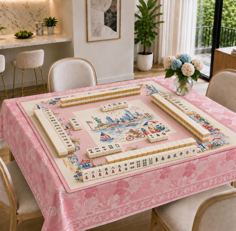 May include: A square mahjong table mat featuring a central illustration of the New York City skyline, the Statue of Liberty, and a bicycle, surrounded by arranged mahjong tiles on a pink tablecloth with a white shell and fan pattern.