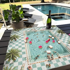 May include: A decorative table mat featuring a pool scene with swimmers, palm trees, and a checkered pattern. The mat is adorned with colorful game pieces. A bottle of champagne and a glass are on the table, suggesting a leisure activity.