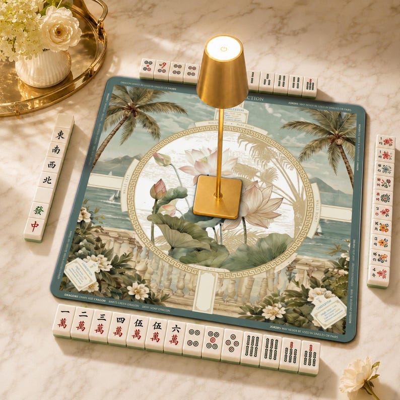 May include: A square mahjong mat featuring a tropical landscape with palm trees and lotus flowers, surrounded by four racks of mahjong tiles with Chinese characters and symbols. A gold-colored metal lamp stands in the center of the mat.