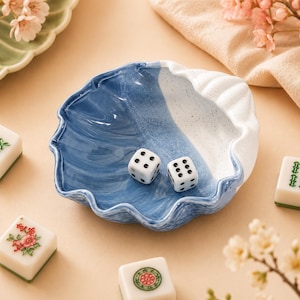 Könnte beinhalten: Eine muschelförmige Keramikschale mit zweifarbigem Design, einer glänzenden blauen Oberfläche auf der einen Seite und einer gesprenkelten weißen Oberfläche auf der anderen. Zwei weiße sechsseitige Würfel mit schwarzen Punkten liegen darin.
