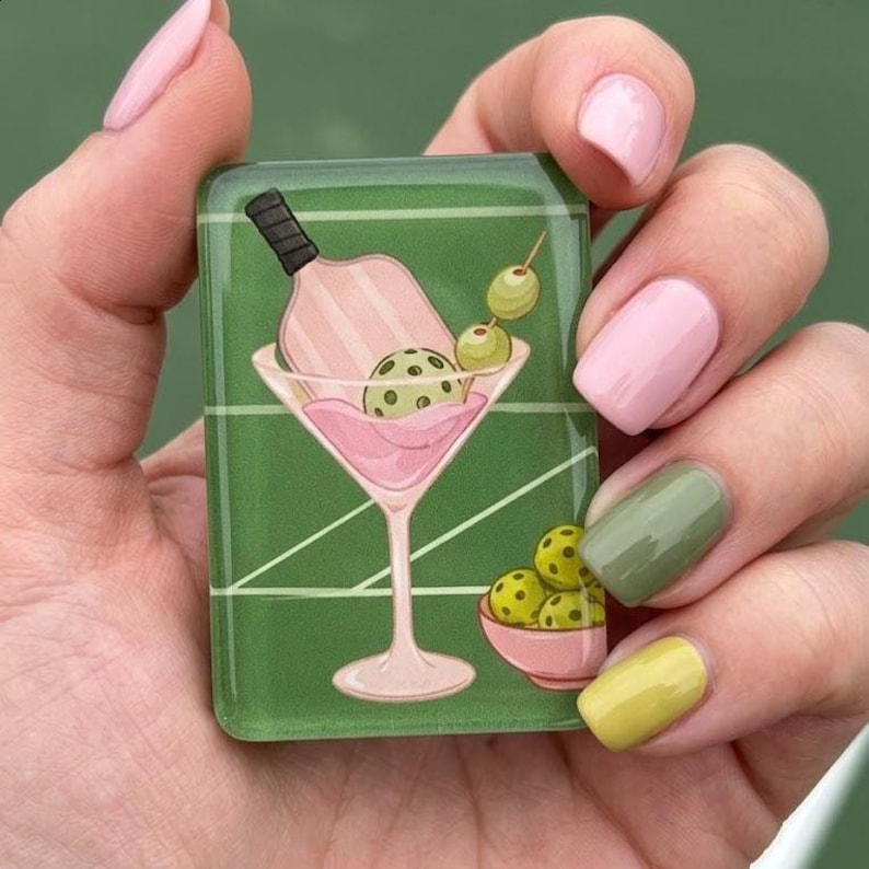 May include: A green rectangular object with a martini glass design. The glass contains a pickleball paddle, a pickleball ball, and olives. A small bowl of pickleballs is also present. The object is held by a hand with pink and green nail polish.