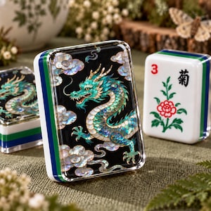 May include: A rectangular mahjong tile featuring a black surface with an iridescent mother-of-pearl dragon design surrounded by clouds. The tile has a white, blue, and green layered side profile. A second tile shows a red number 3 and a flower.