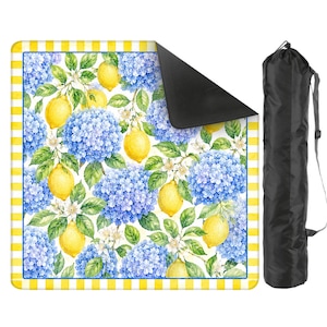 May include: A picnic blanket featuring a vibrant watercolor design of lemons, blue hydrangeas, and white flowers on a white background. The blanket has a yellow and white checkered border and a black underside. A black carrying bag is included.