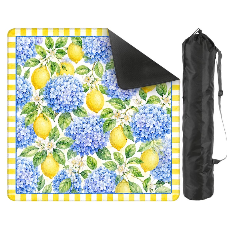 May include: A picnic blanket featuring a vibrant watercolor design of lemons, blue hydrangeas, and white flowers on a white background. The blanket has a yellow and white checkered border and a black underside. A black carrying bag is included.