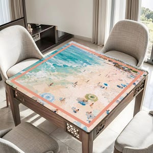 May include: A square wooden table featuring a beach scene print. The top depicts a sandy beach with figures, parasols, and the sea. The table has a decorative border and is surrounded by chairs.