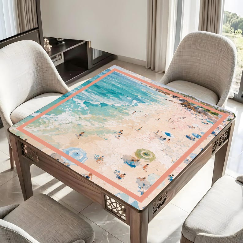 May include: A square wooden table featuring a beach scene print. The top depicts a sandy beach with figures, parasols, and the sea. The table has a decorative border and is surrounded by chairs.