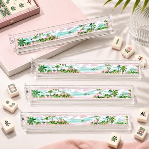 May include: Clear acrylic mahjong tile holders with a tropical landscape design. The holders feature a watercolor scene of palm trees, ocean, and sky in shades of green, blue, and pink. Several mahjong tiles are scattered around the holders.