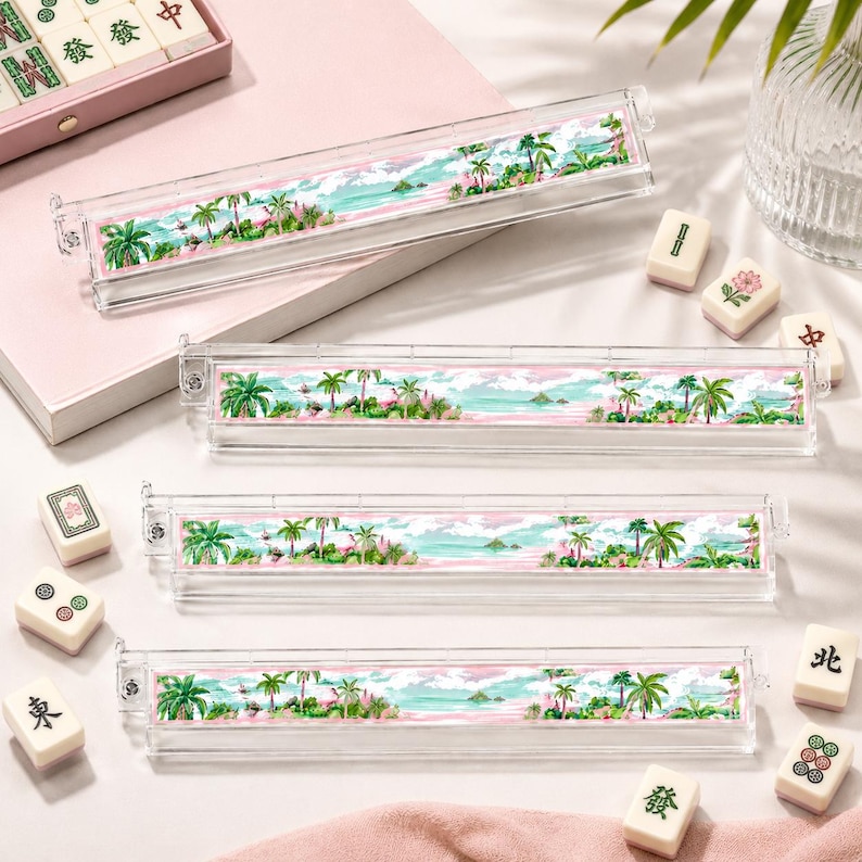 May include: Clear acrylic mahjong tile holders with a tropical landscape design. The holders feature a watercolor scene of palm trees, ocean, and sky in shades of green, blue, and pink. Several mahjong tiles are scattered around the holders.