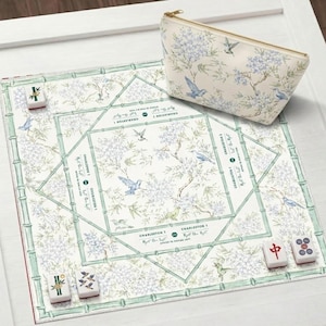 Op de afbeelding: Een vierkante mahjongmat met een witte achtergrond, lichtgroene bamboeranden en blauwe bloemen- en vogelpatronen. De mat bevat de gedrukte tekst Charleston 1. Een bijpassend tasje met rits en vier mahjongstenen zijn inbegrepen.