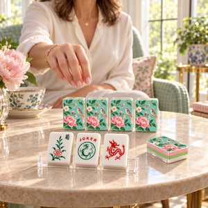 May include: A set of rectangular mahjong tiles featuring a light blue background with pink floral and bird illustrations. Three tiles are displayed on a clear acrylic stand, showing a flower, a green dragon, and a red dragon with the text JOKER.