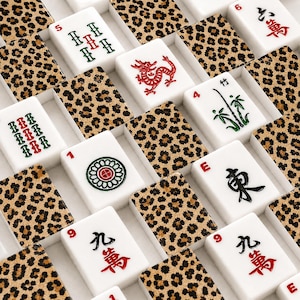 Puede incluir: Primer plano de un juego de Mahjong con fichas blancas y detalles de estampado de leopardo. Las fichas presentan varios s&iacute;mbolos, n&uacute;meros y caracteres chinos en rojo, verde y negro. El juego est&aacute; dispuesto en forma de cuadr&iacute;cula.