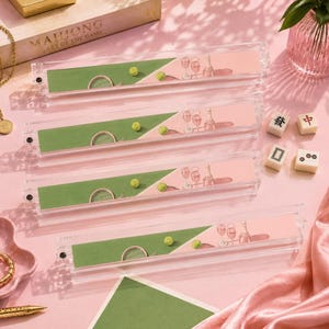 May include: Four clear acrylic Mahjong tile holders with pink and green decorative inserts. Each holder features a tennis court design with a green court, a yellow tennis ball, and champagne glasses. The game box is visible in the background.