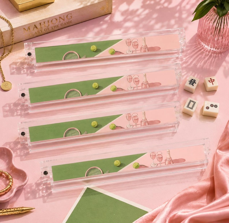 May include: Four clear acrylic Mahjong tile holders with pink and green decorative inserts. Each holder features a tennis court design with a green court, a yellow tennis ball, and champagne glasses. The game box is visible in the background.