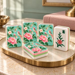 May include: Four rectangular mahjong tiles with a teal background featuring pink peonies and small birds, plus one white tile with a green and red floral design and a black character. The tiles are arranged on an oval gold-rimmed tray.