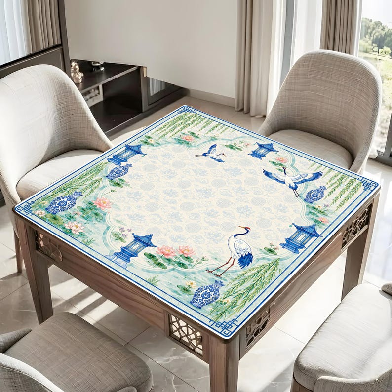 May include: A square tablecloth featuring a white centre with a subtle circular pattern, surrounded by a border of blue pagodas, white and blue vases, pink lotus flowers, green willow branches, and white cranes with black and red accents.