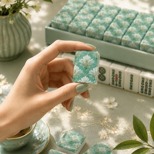 May include: A close-up of a teal and white Mahjong tile with a floral design, held between fingers. Other tiles with the same design are in a tray, and more are visible in the background. A cup of tea and a vase of flowers are also in the image.