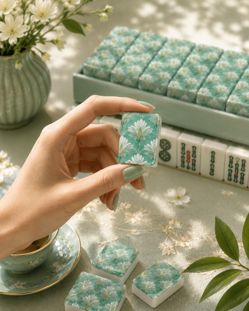 May include: A close-up of a teal and white Mahjong tile with a floral design, held between fingers. Other tiles with the same design are in a tray, and more are visible in the background. A cup of tea and a vase of flowers are also in the image.