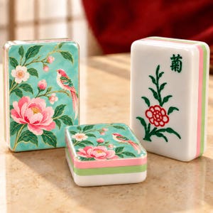 May include: Three rectangular mahjong tiles with rounded edges. Two tiles feature a teal background with pink flowers, green leaves, and a small bird. One white tile displays a green plant, a red flower, and the black Chinese character for chrysanthemum.