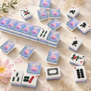 Op de afbeelding: Een set rechthoekige mahjongtegels met een witte basis met zwarte, rode en groene inkttekens of symbolen, en een blauwachtig bovenoppervlak versierd met een roze waterlelie-illustratie. Sommige tegels zijn in een rij gerangschikt.