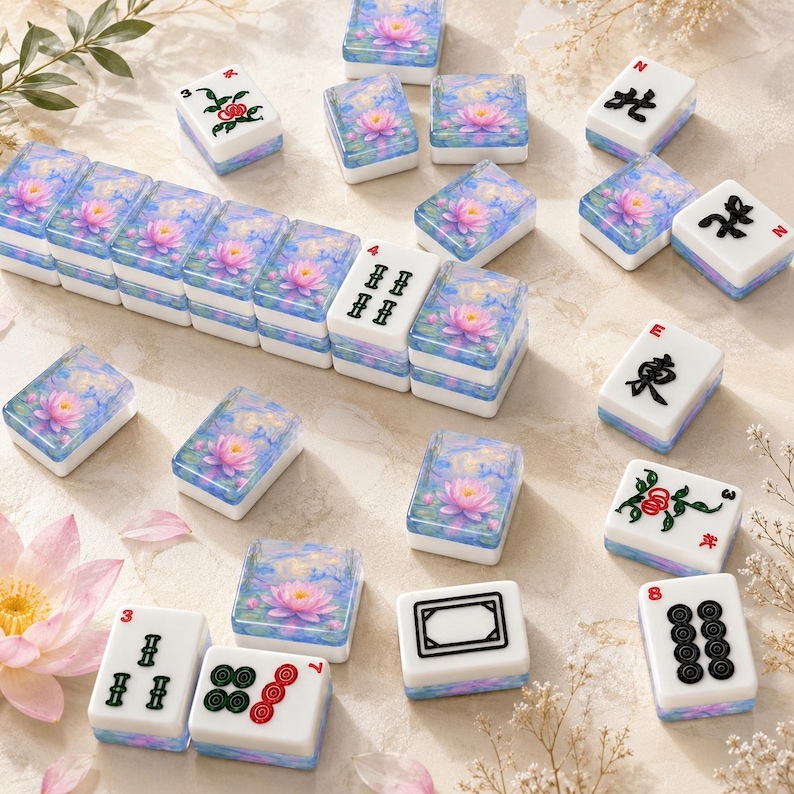 Op de afbeelding: Een set rechthoekige mahjongtegels met een witte basis met zwarte, rode en groene inkttekens of symbolen, en een blauwachtig bovenoppervlak versierd met een roze waterlelie-illustratie. Sommige tegels zijn in een rij gerangschikt.