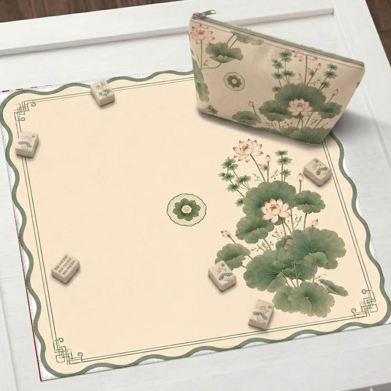 May include: A square game mat with a scalloped edge and a cream background featuring a green lotus flower and leaf pattern, accompanied by a matching zippered pouch and several small rectangular game tiles scattered across the surface.
