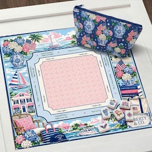 May include: A blue and white patterned cosmetic bag and a matching game board featuring coastal imagery, sailboats, and American flags. The board has a pink center and text that reads "Charleston" and "Hamptons Living."