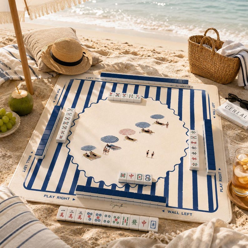 May include: A beach-themed mahjong set with a blue and white striped mat. The mat features a scalloped center with illustrations of beach umbrellas and people. The set includes white tiles, a straw hat, and a woven basket on a sandy beach.