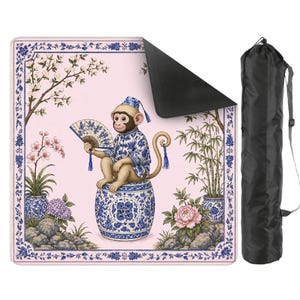 May include: A pink yoga mat featuring a monkey in blue and white traditional clothing, holding a fan. The mat has a floral and botanical design with a black underside. A black carrying bag is included.