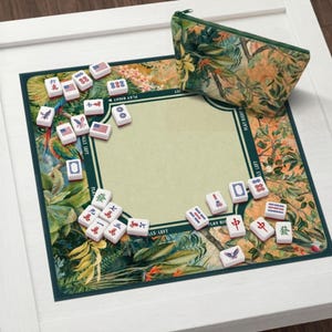 Pode incluir: Conjunto de jogo de mahjong com um design floral tropical. O conjunto inclui um tabuleiro de jogo verde e bege, peças e uma bolsa com zíper a condizer. O tabuleiro apresenta uma área de jogo central e uma borda com texto. As peças têm vários símbolos e números.