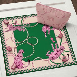 Könnte beinhalten: Eine quadratische Matte mit einer einfarbigen grünen Mitte, einem hellrosa Rand mit dunkelgrün-rosa Karomuster sowie rosa Leoparden-Illustrationen und Kettenmotiven. Eine passende rosa Reißverschlusstasche mit Leoparden- und Kettenmuster liegt bei.